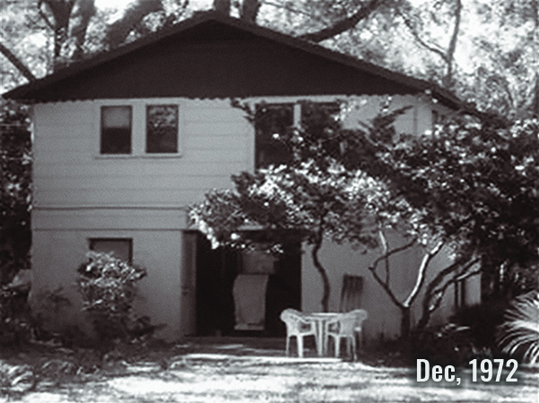 Aged photo of a house with open garage door and plastic chairs in front.